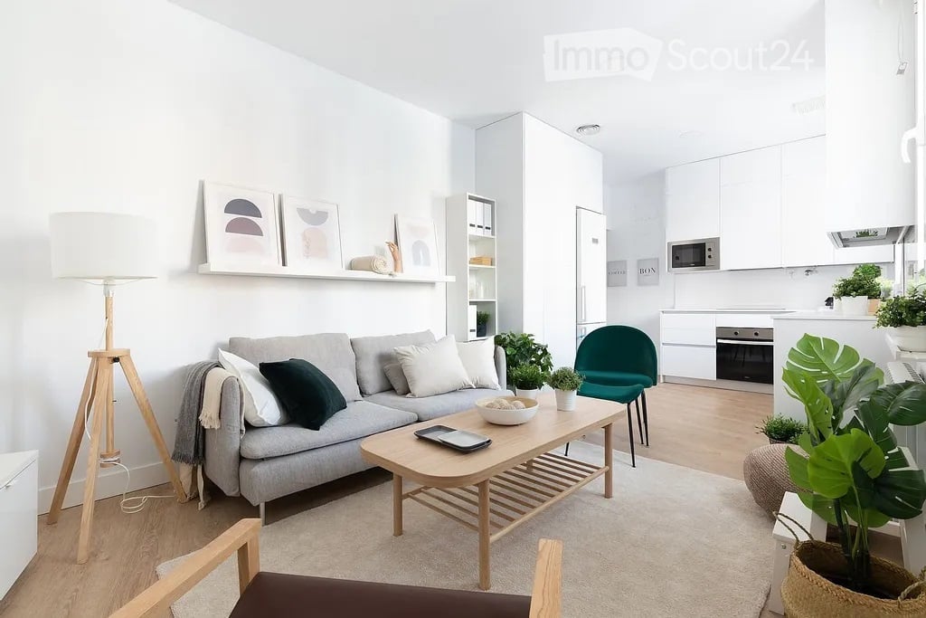 Modern living room with a grey sofa, wooden coffee table, floor lamp, green accent chair, and white kitchen area with microwave and oven. Shelves with decorative items and potted plants are present.