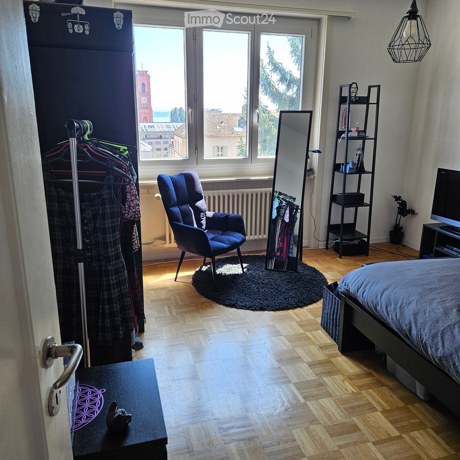 Well-lit bedroom with wooden flooring, large windows offering a city view, a bed with blue bedding, a blue armchair, a mirror, a shelf, a small table with a decorative item, and a clothing rack with hanging clothes.
