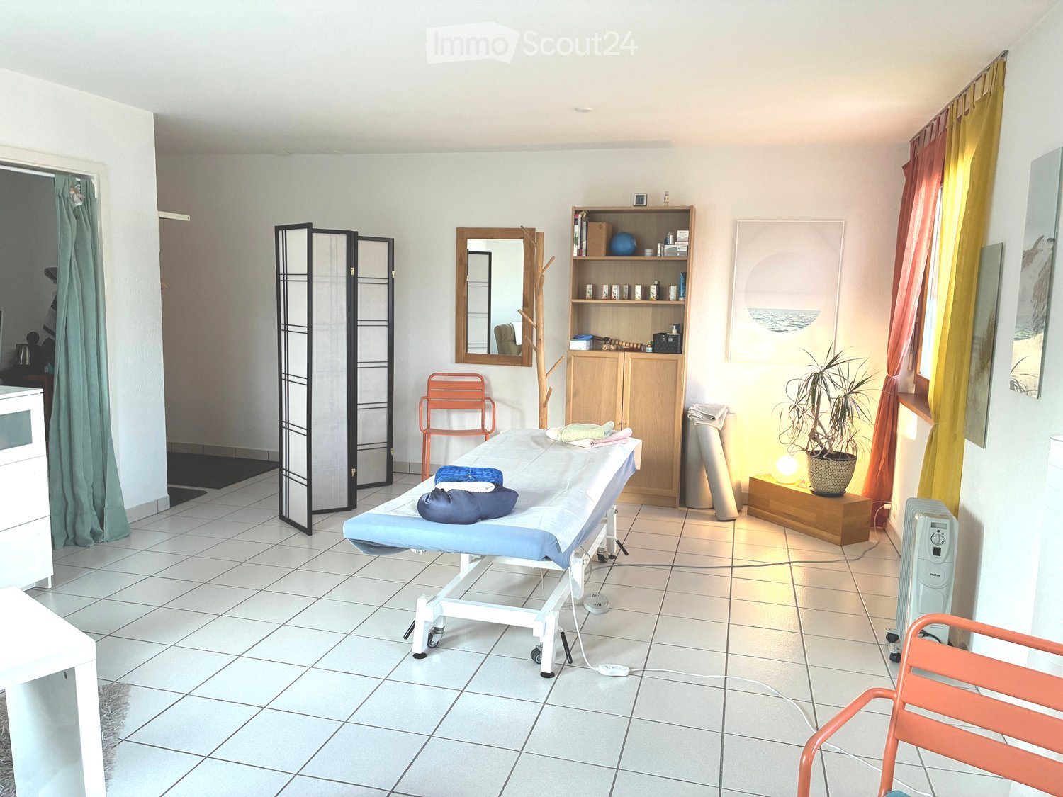 A well-lit room with a massage table, a wooden cabinet, a mirror, a potted plant, and a screen. The room features tiled flooring and has a cozy ambiance with warm lighting.