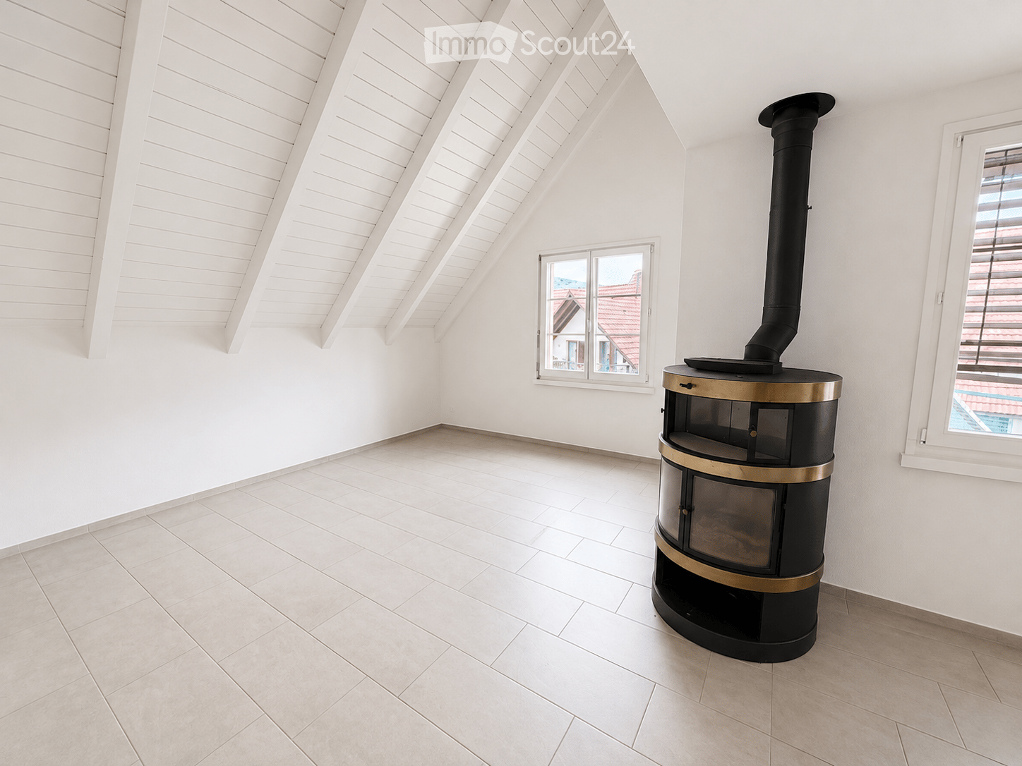 Empty room with white walls, light-colored tiled floor, and a modern wood-burning stove with a chimney. Two windows offer views of the outside.