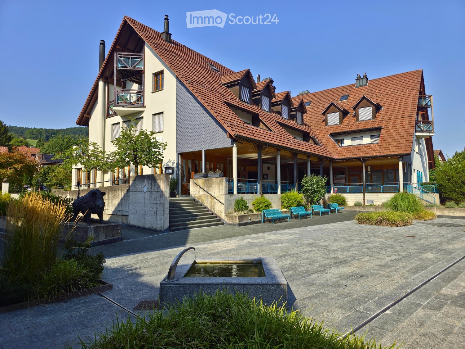 A modern building with a red-tiled roof, multiple balconies, and a covered entrance area. The surrounding area includes a paved courtyard with benches, a small water feature, and landscaped greenery.