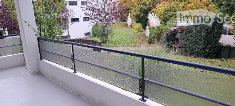Balcony with glass railing, overlooking a green lawn and trees, with a building in the background.