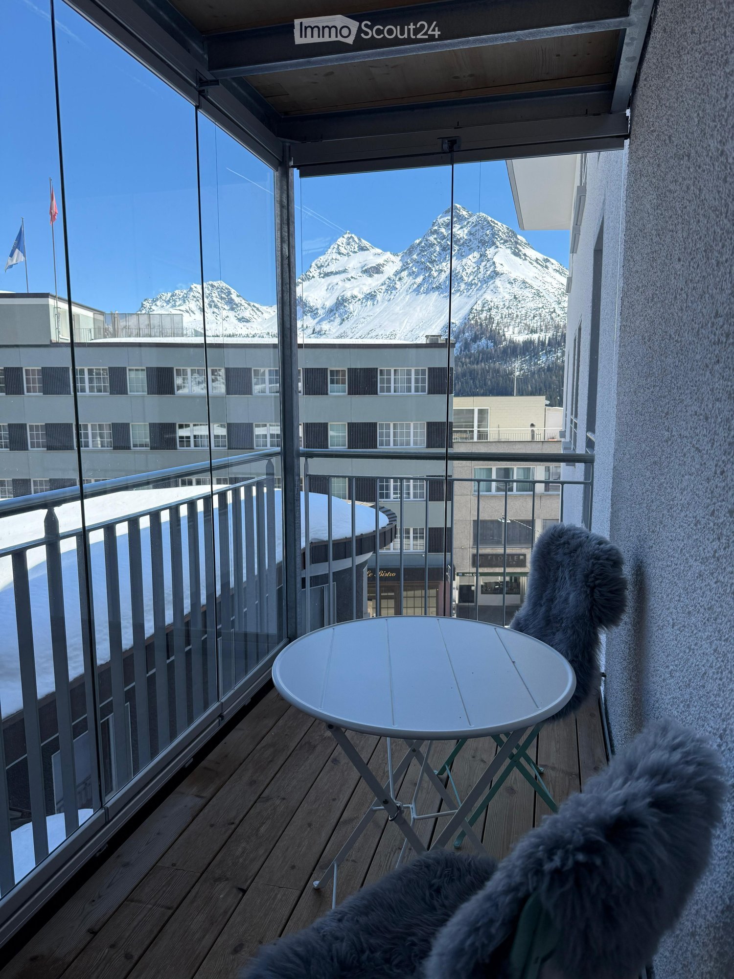 Balcony with wooden decking, white round table, and a fur-covered chair, overlooking a snow-capped mountain and urban buildings.