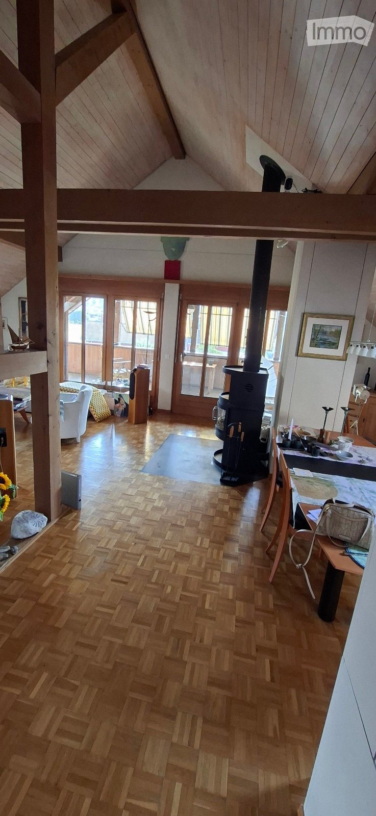 Spacious living room with wooden herringbone flooring, white walls, and exposed wooden beams. Features a wood stove, sofa, chair, and a small table with items on it.