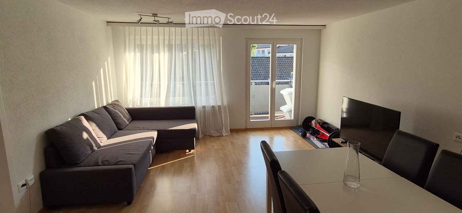 Bright living room with wooden flooring, a grey sectional sofa, a dining table with chairs, and a television. A sliding glass door leads to a balcony.