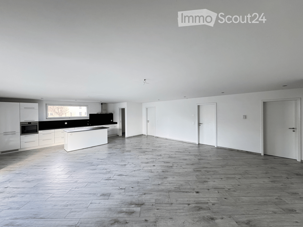 Open-plan space with a modern kitchen area featuring white cabinets, a central island, and a black countertop. The main area has a wooden floor and white walls, with several doors leading to other rooms.