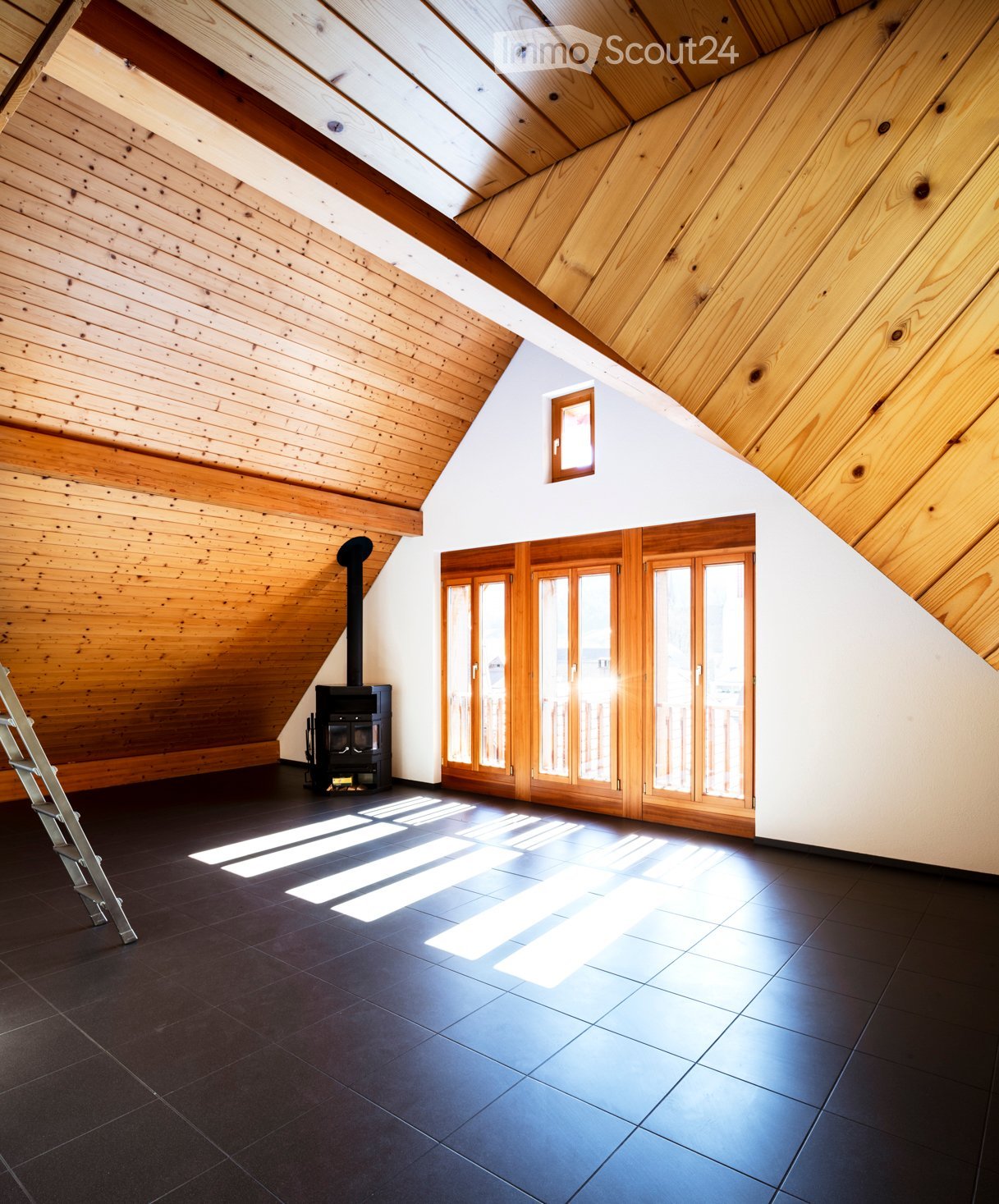 An attic space with wooden slanted ceilings, dark tiled floor, and large wooden-framed windows. A black wood-burning stove is positioned against the wall, and a ladder is visible on the left side.