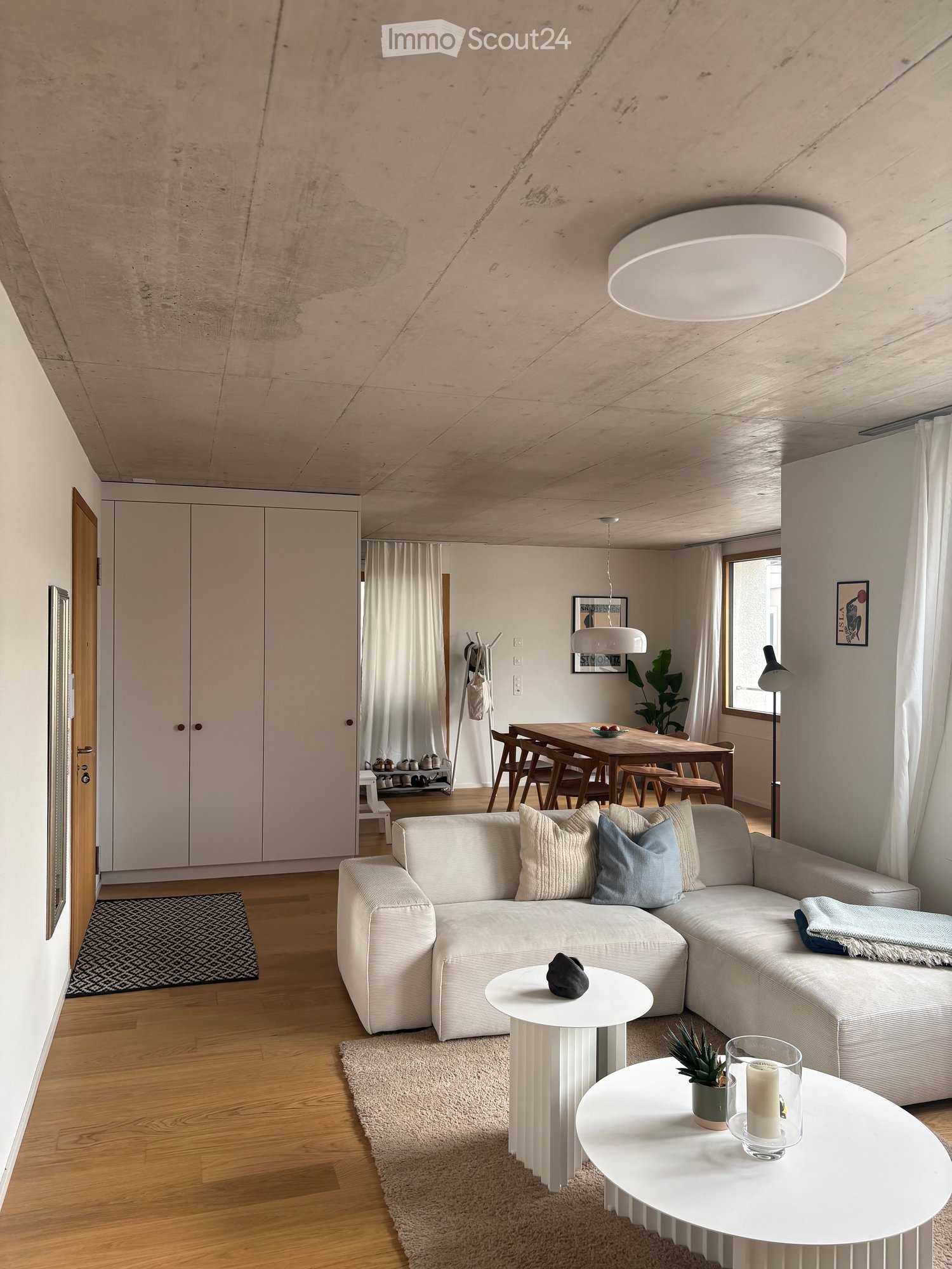 Modern living room with a white sectional sofa, coffee tables, dining area with a wooden table and chairs, and a sleek cabinet. The space features wooden flooring, large windows with white curtains, and contemporary lighting.