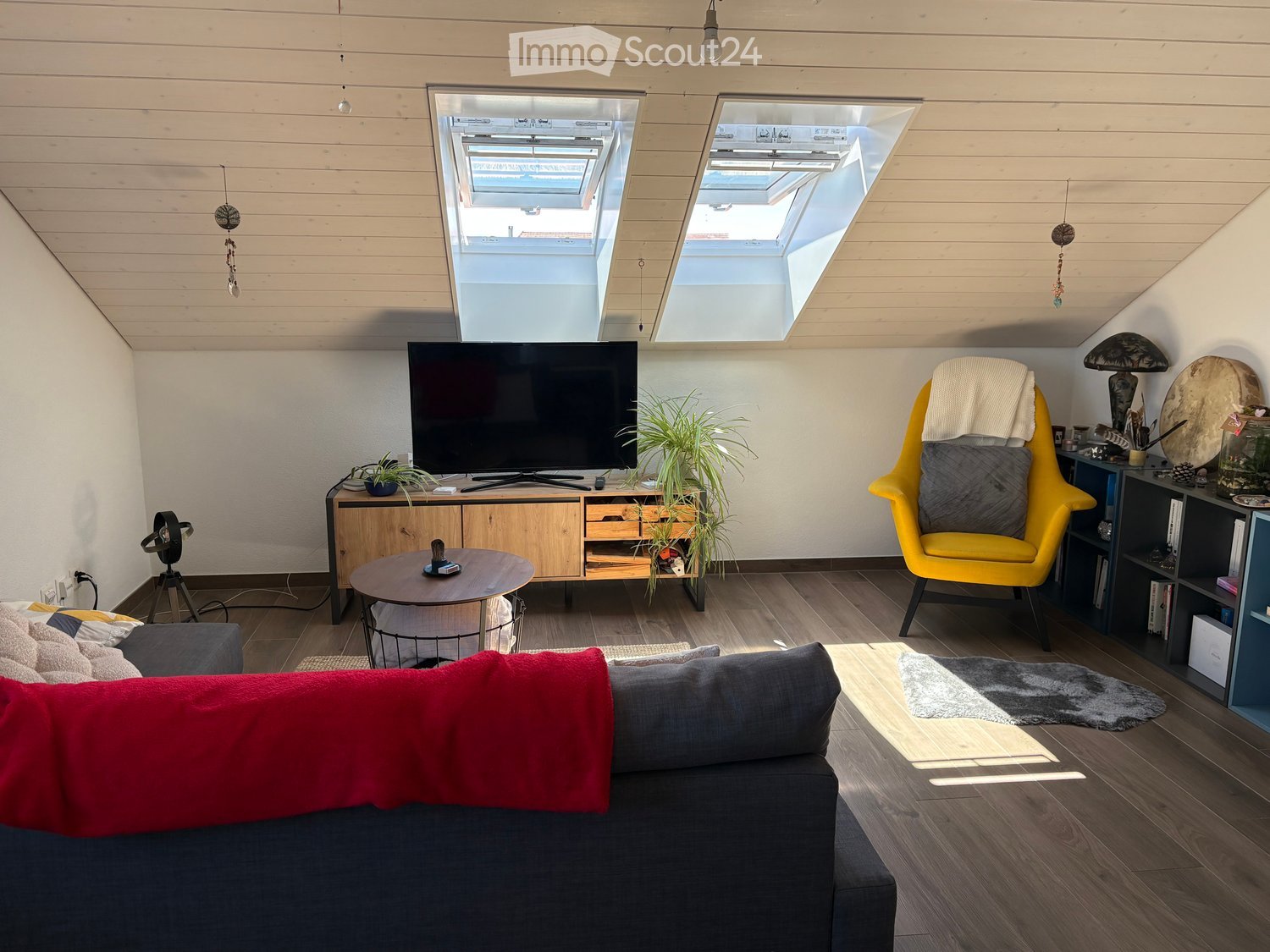 Modern living room with wooden floors, slanted ceiling, skylights, grey sofa, coffee table, yellow armchair, TV, and decorative plants.