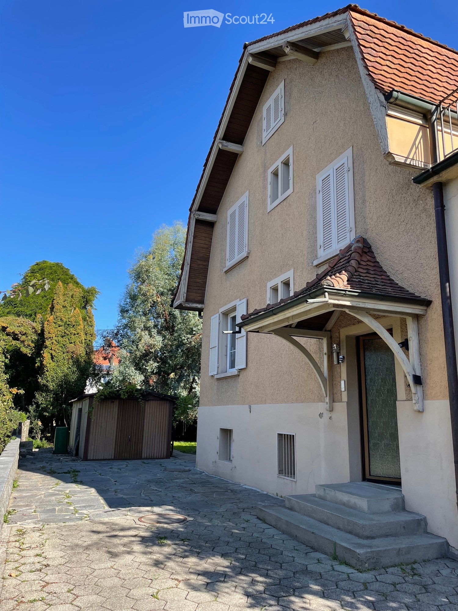 Two-story house with beige facade, multiple windows with shutters, and a small porch with wooden beams. A stone pathway leads to the entrance.