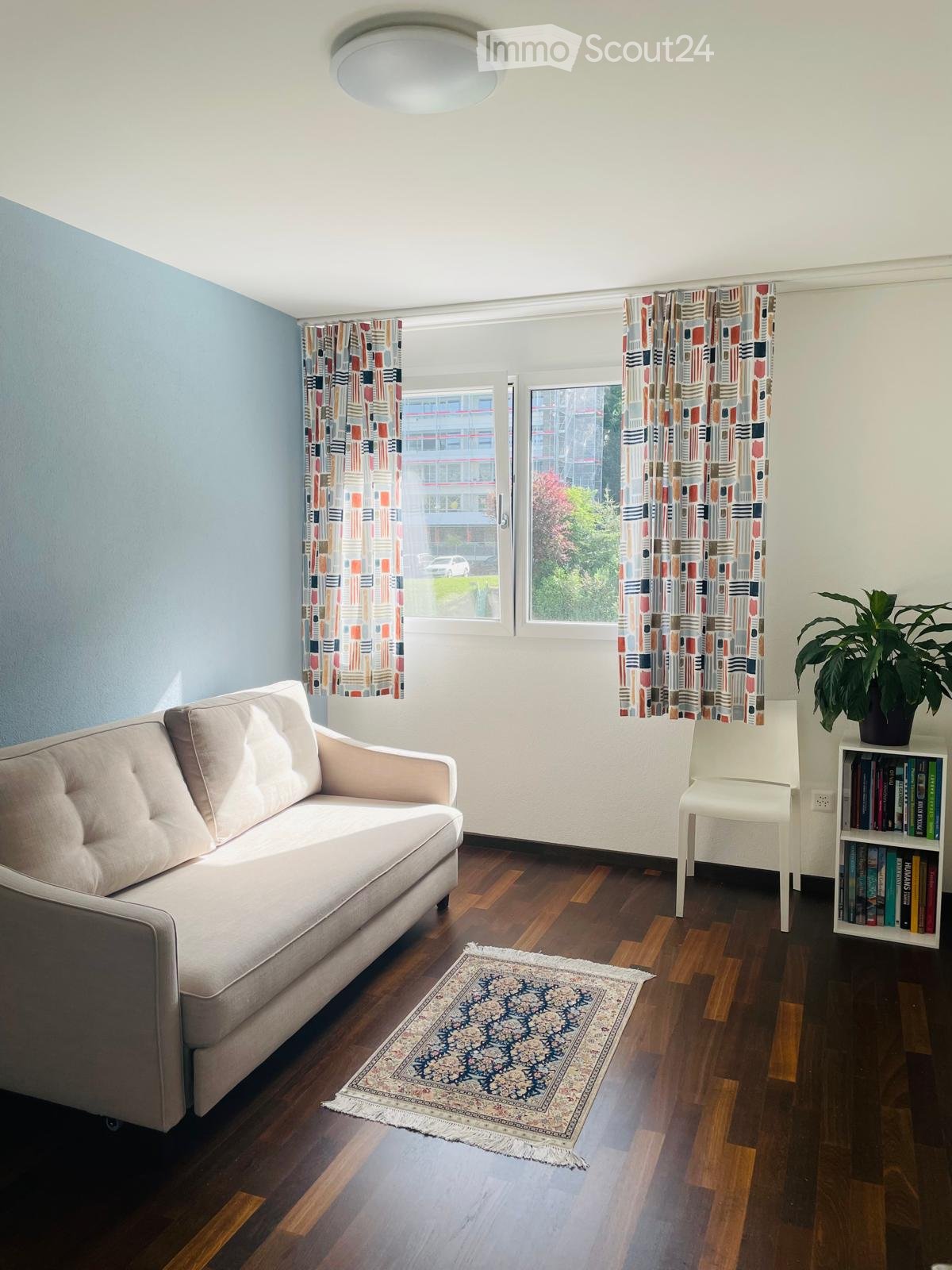 Living room with a light blue wall, wooden floor, and large windows with colorful curtains. Features a beige sofa, white chair, bookshelf with books, a potted plant, and a patterned rug.