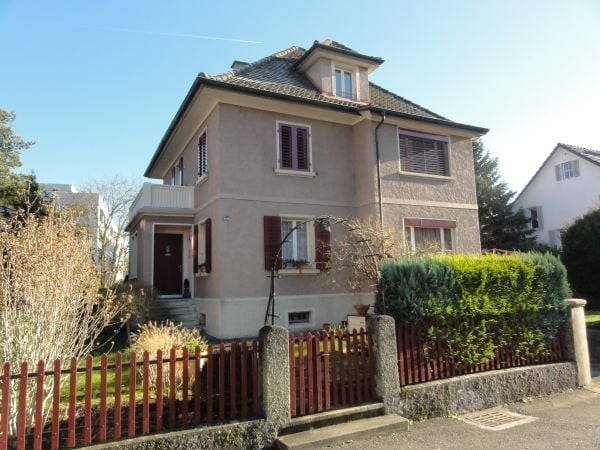Two-story house with beige facade, multiple windows, and a small balcony on the upper floor. The house is surrounded by a low fence and some greenery.