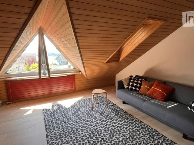 Modern living room with wooden ceiling, large triangular window, grey sofa with patterned cushions, small wooden stool, and a patterned rug.