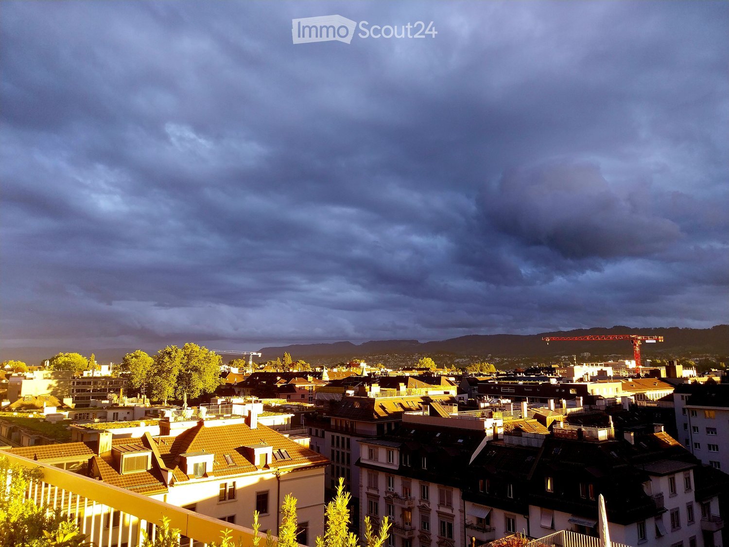 Panoramic cityscape view with rooftops, trees, and distant mountains under a dramatic sky.