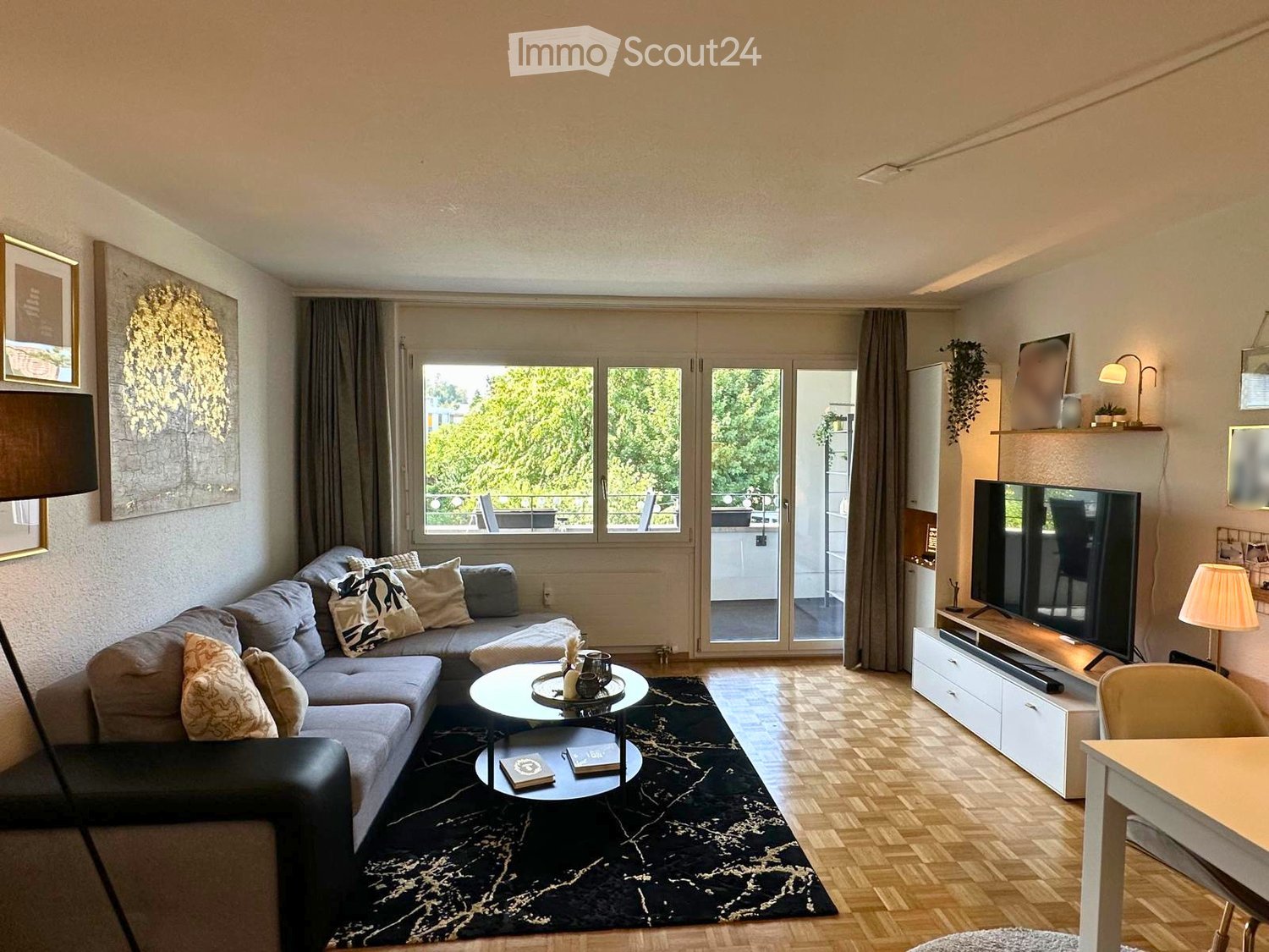 Modern living room with grey sectional sofa, coffee table, TV, and large windows offering a view of greenery.