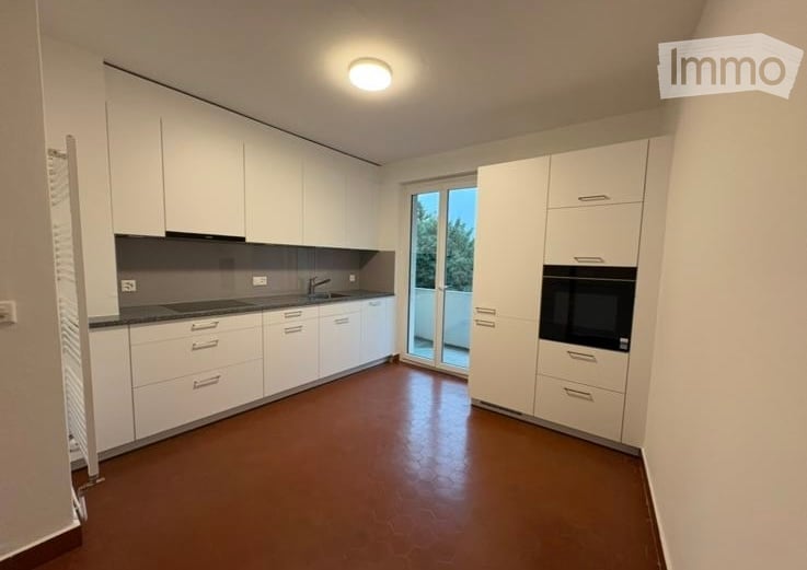 Modern, minimalist kitchen with white cabinets, a dark countertop, and a built-in oven. Features a large window with a sliding glass door leading to a balcony.