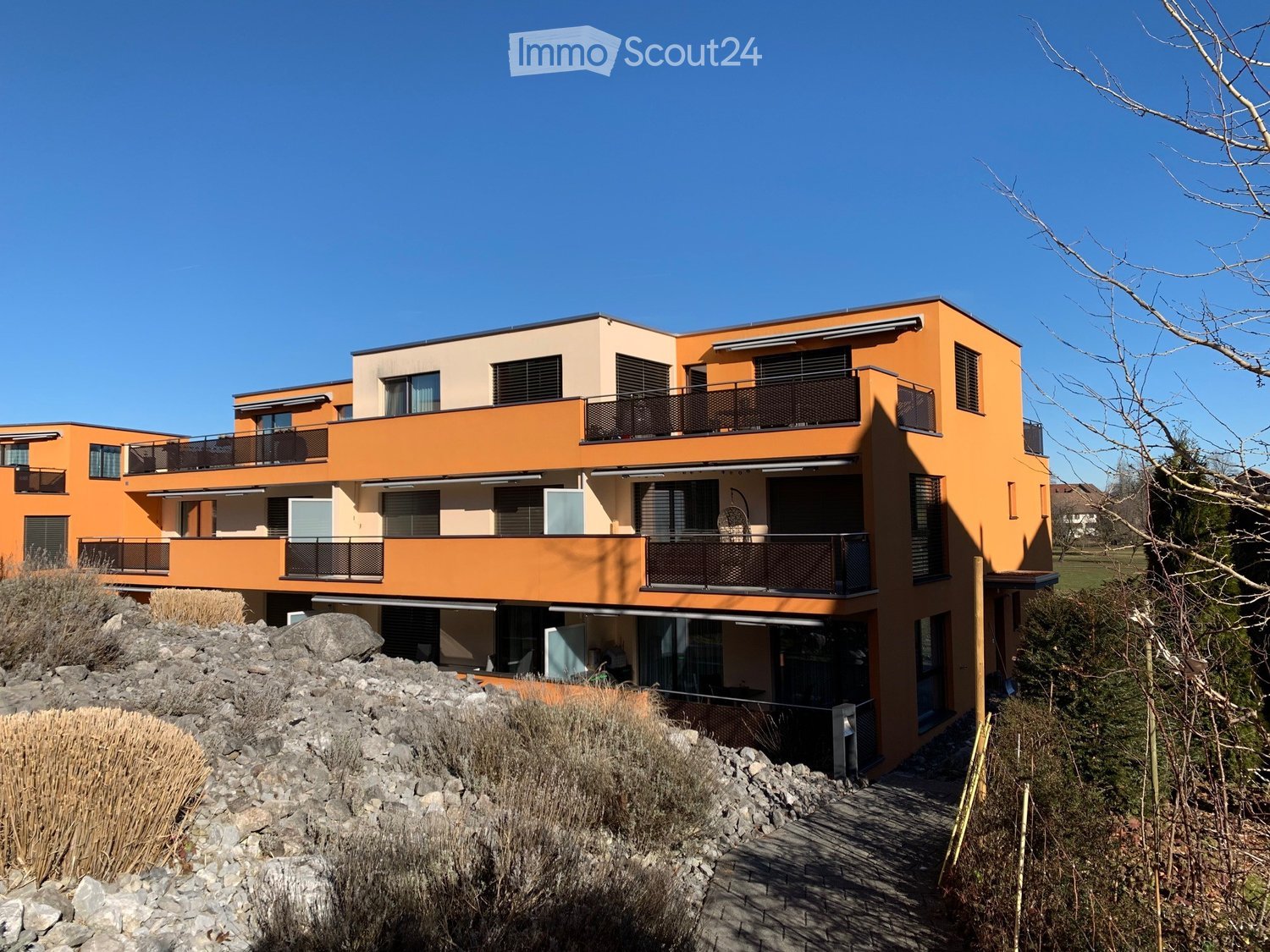 Modern two-story house with orange and beige exterior, multiple balconies, large windows, and a raised ground floor.