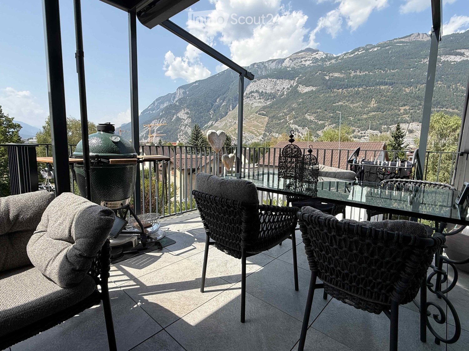 Balcony with a view of mountains, featuring a sofa, chairs, a table, and a grill.