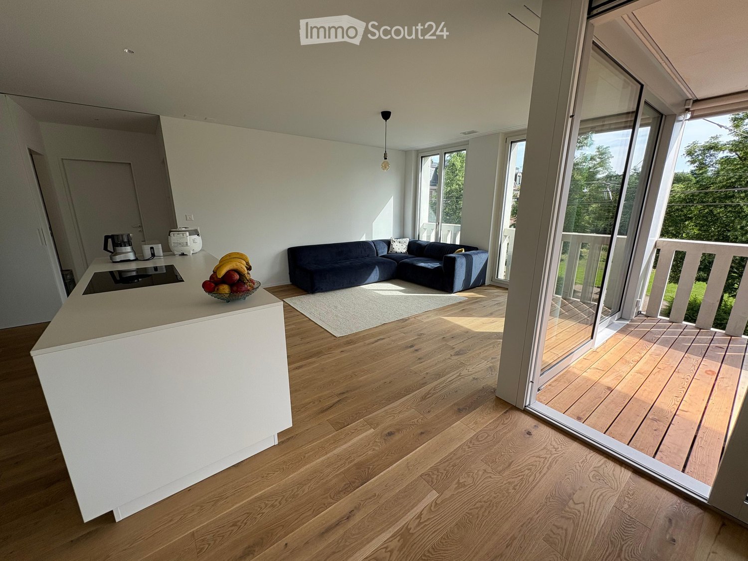 Modern living room with a kitchen island, dark blue sectional sofa, and large windows opening to a balcony. The room features light wooden flooring and a white rug.