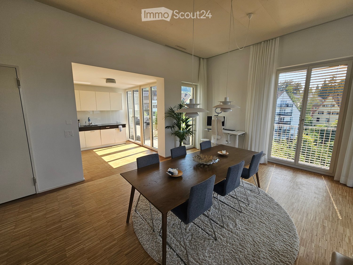 Open-plan dining and kitchen area with wooden flooring, large windows, and modern furniture including a dining table, chairs, pendant lights, and a small kitchen with cabinets and a sink.