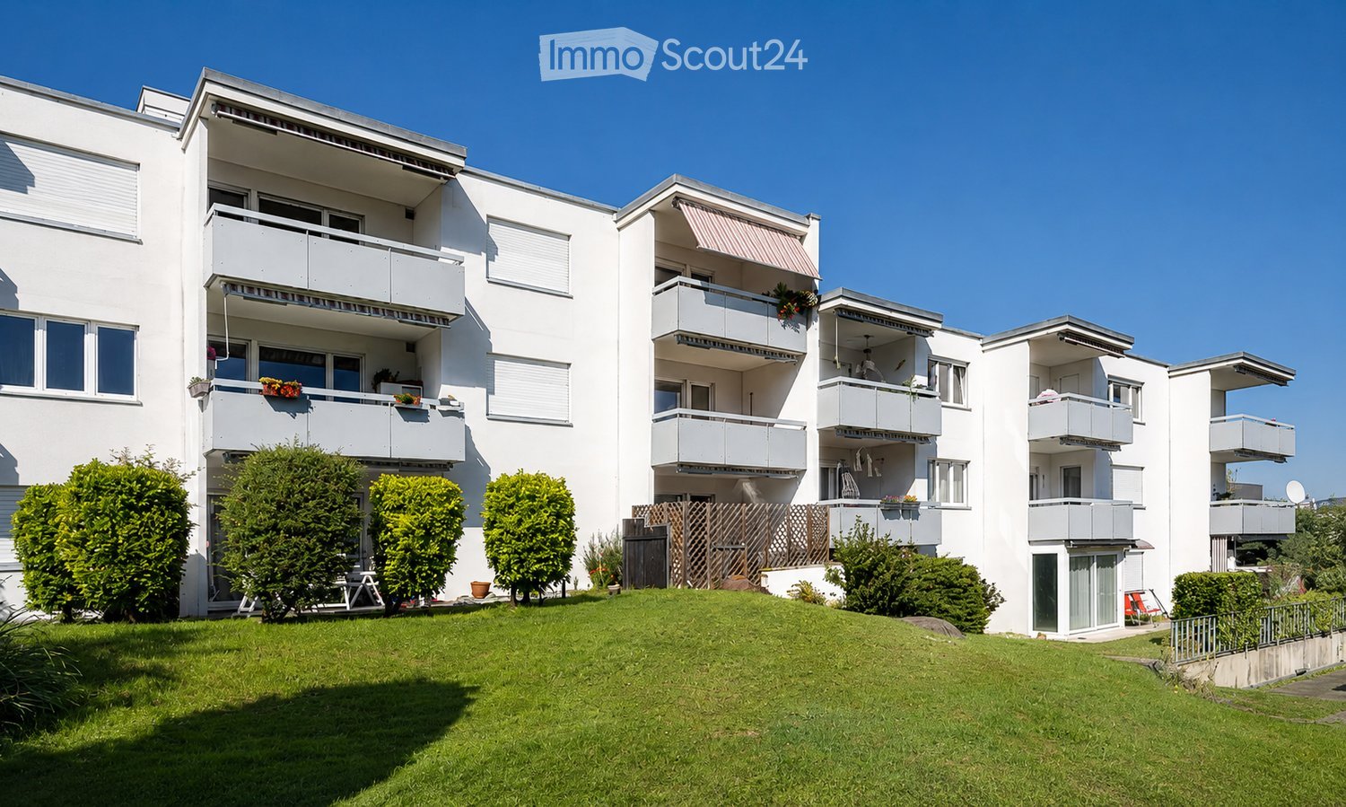 Modern white apartment building with multiple balconies, greenery, and a well-maintained lawn in front.