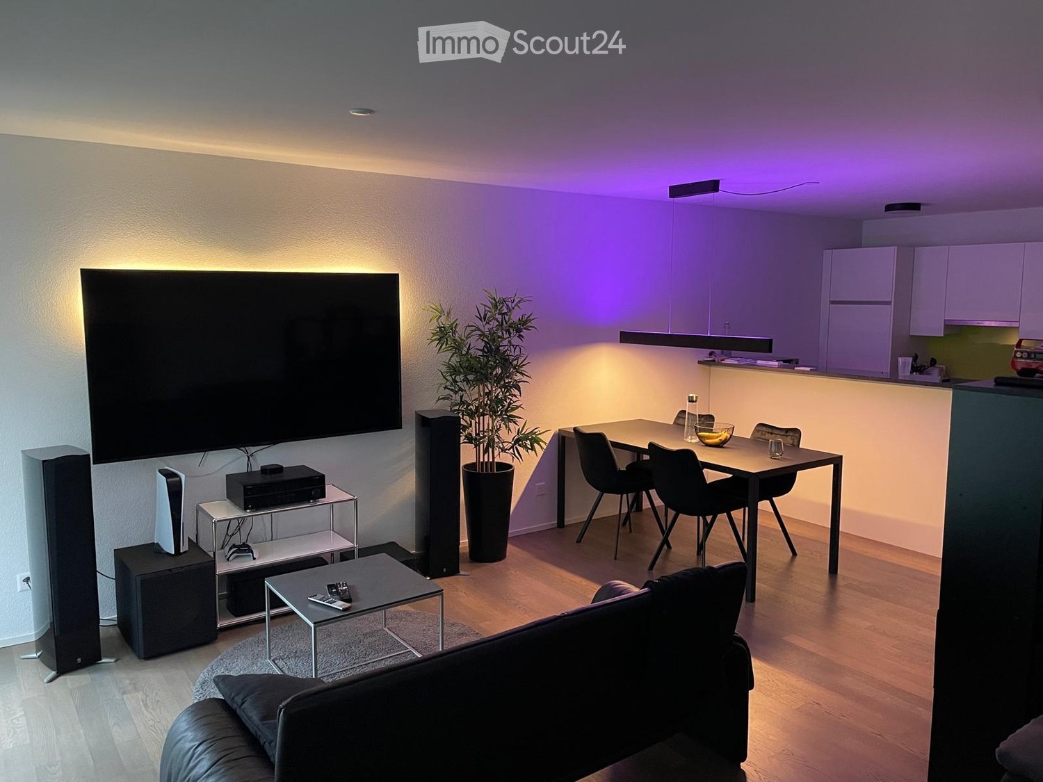 Modern living room with a large screen, sofa, coffee table, dining table with chairs, and a potted plant. The room features wooden flooring and ambient lighting.
