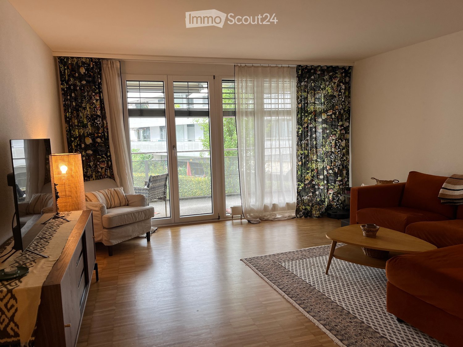 Modern living room with wooden flooring, large sliding glass doors leading to a balcony, a red sofa, a light-colored armchair, a coffee table, a TV stand, and a floor lamp.