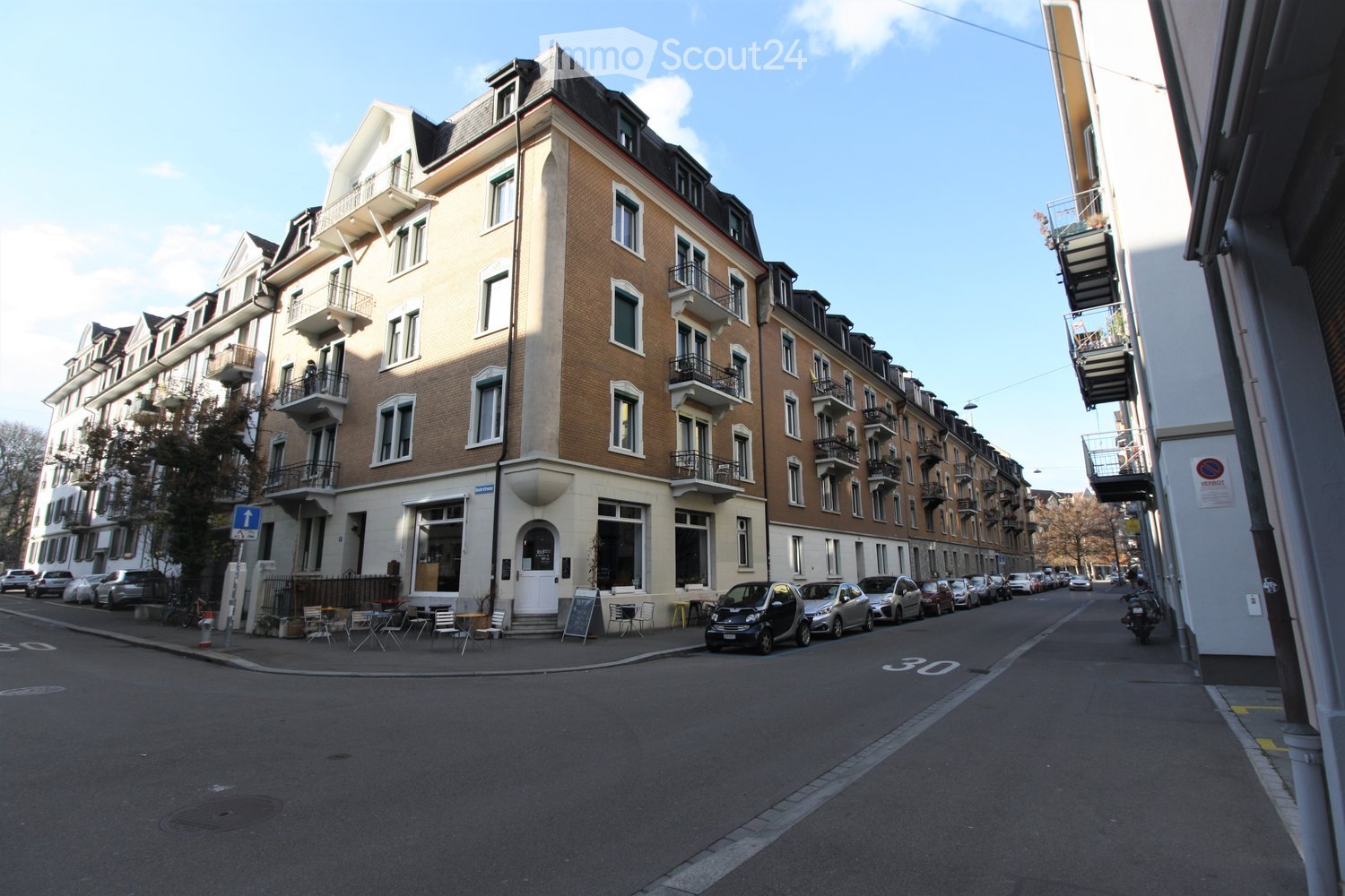 Multi-story brick building with balconies and large windows, located on a quiet street with parked cars and outdoor seating area.