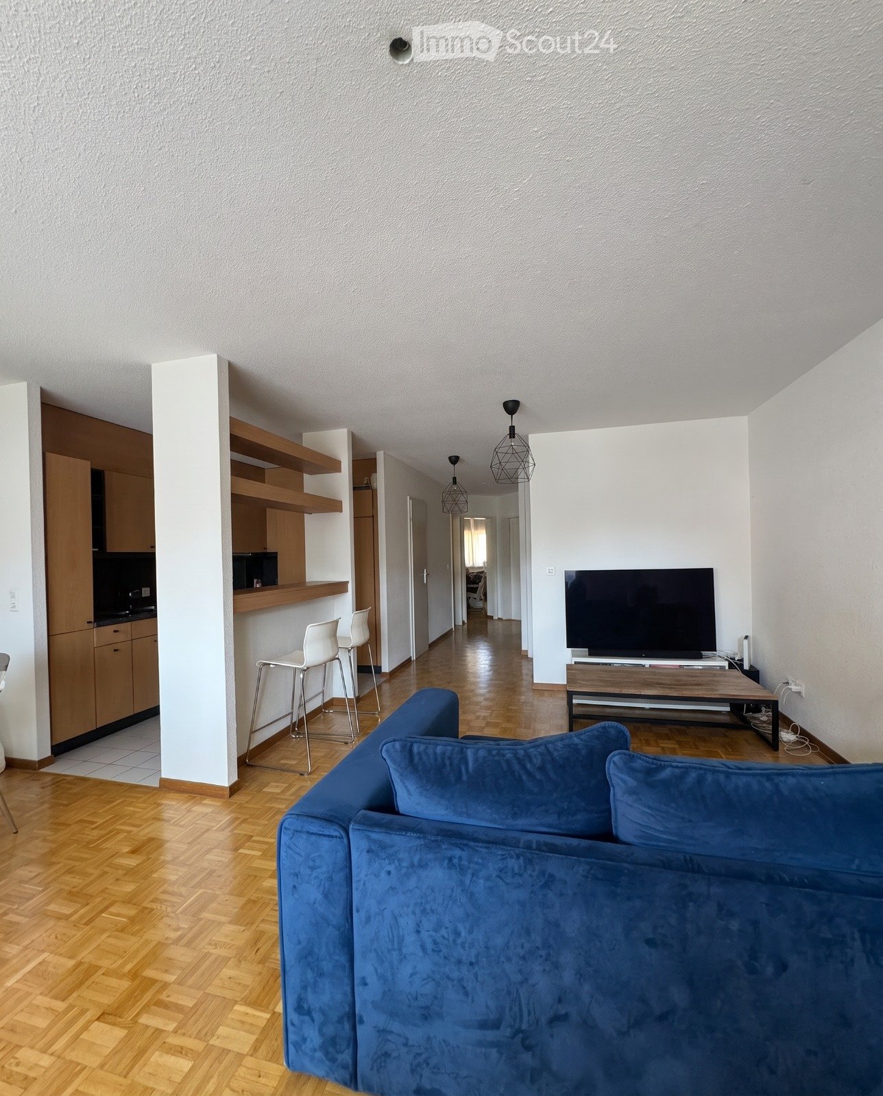 Modern living room with a blue sofa, wooden coffee table, TV, and two white stools near the kitchen area.