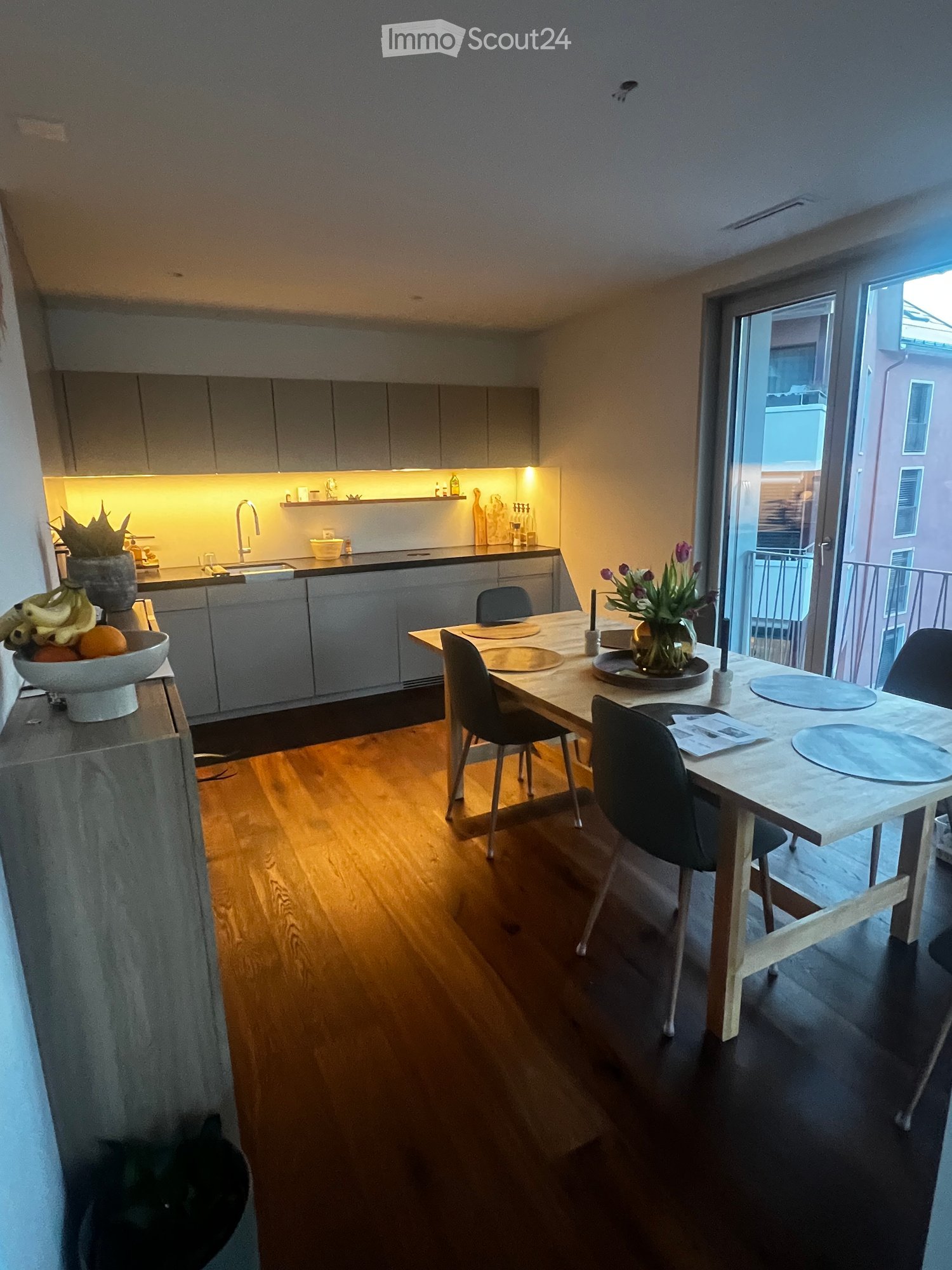 Modern kitchen and dining area with wooden flooring, grey cabinets, a dining table with chairs, and a vase of flowers on the table.