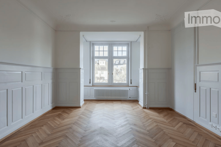 Empty room with wooden herringbone flooring, white walls, large window with radiator below, and white paneling on walls.