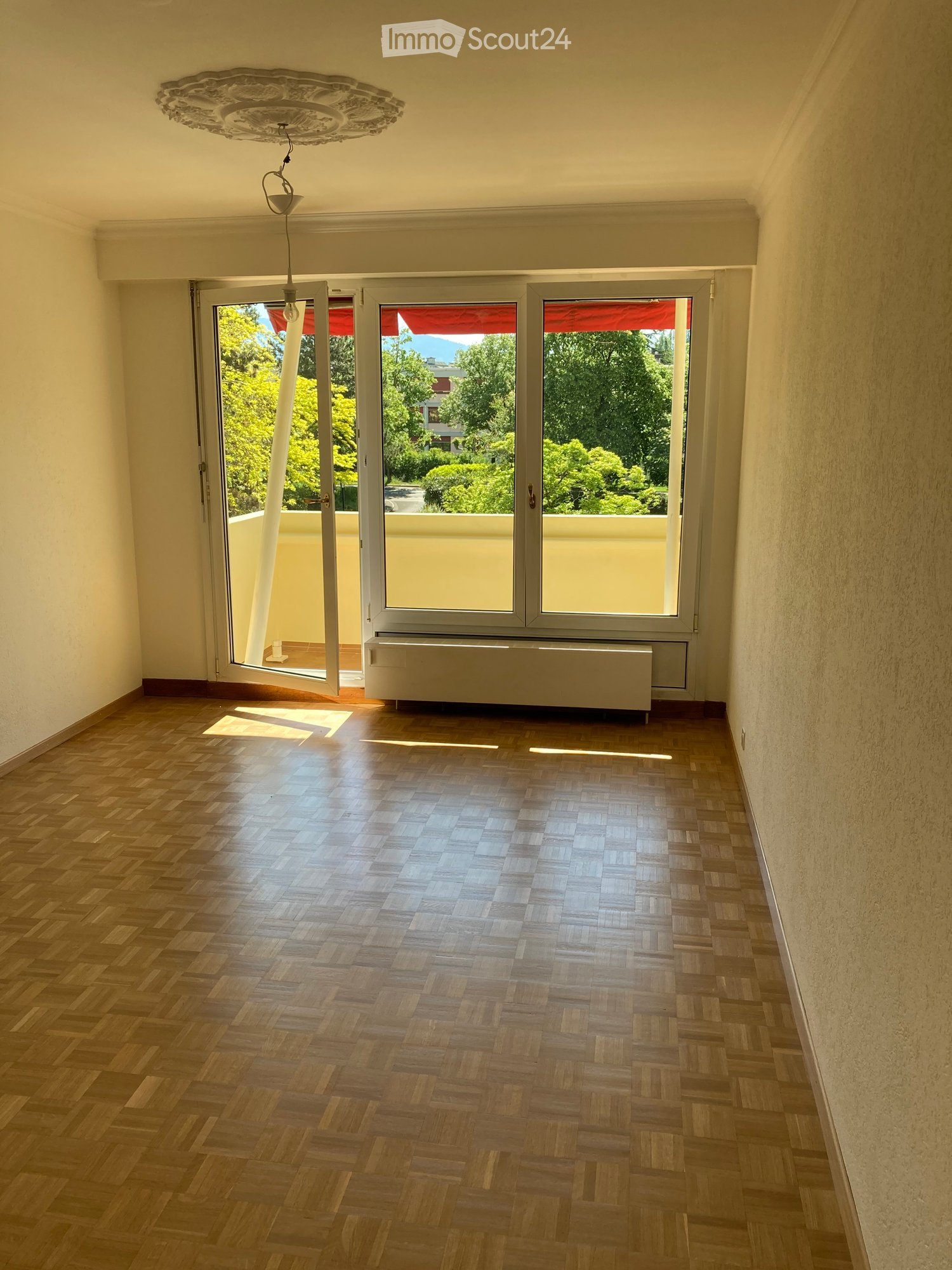 Empty room with wooden herringbone flooring, large windows with red blinds, and a hanging light fixture.