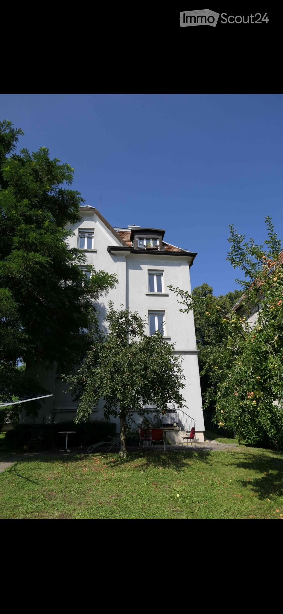 A two-story white house with a pitched roof, surrounded by a well-maintained garden with fruit trees and outdoor seating.