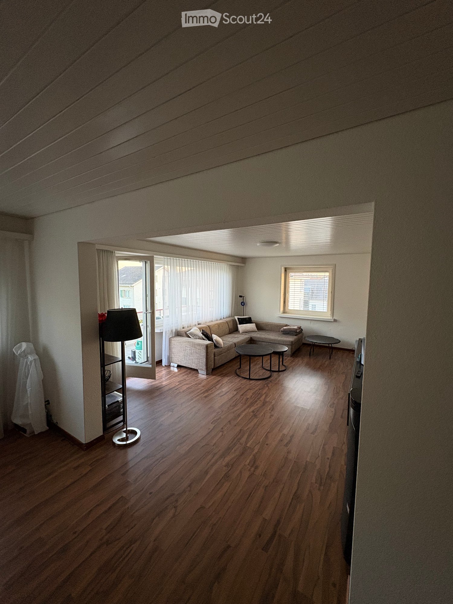 Bright living room with wooden flooring, a sectional sofa, coffee table, side tables, and a floor lamp. Large windows and an open door lead to a balcony.