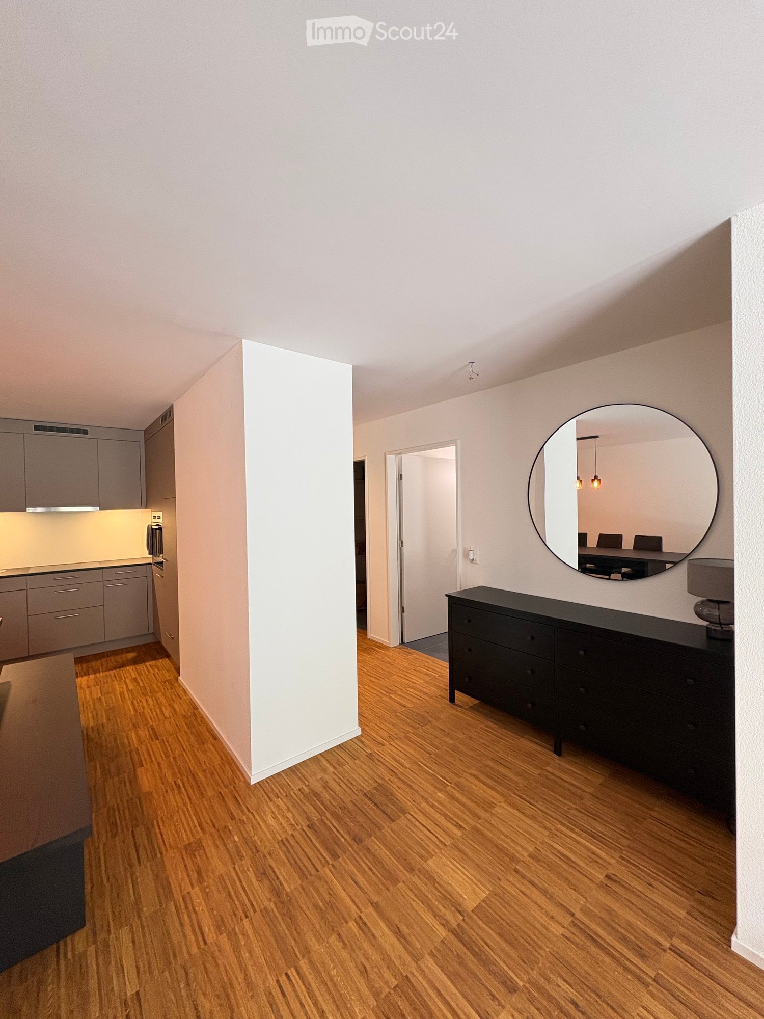 A modern, well-lit hallway with wooden flooring, a black cabinet, a large round mirror, and a small table with chairs visible in the reflection.