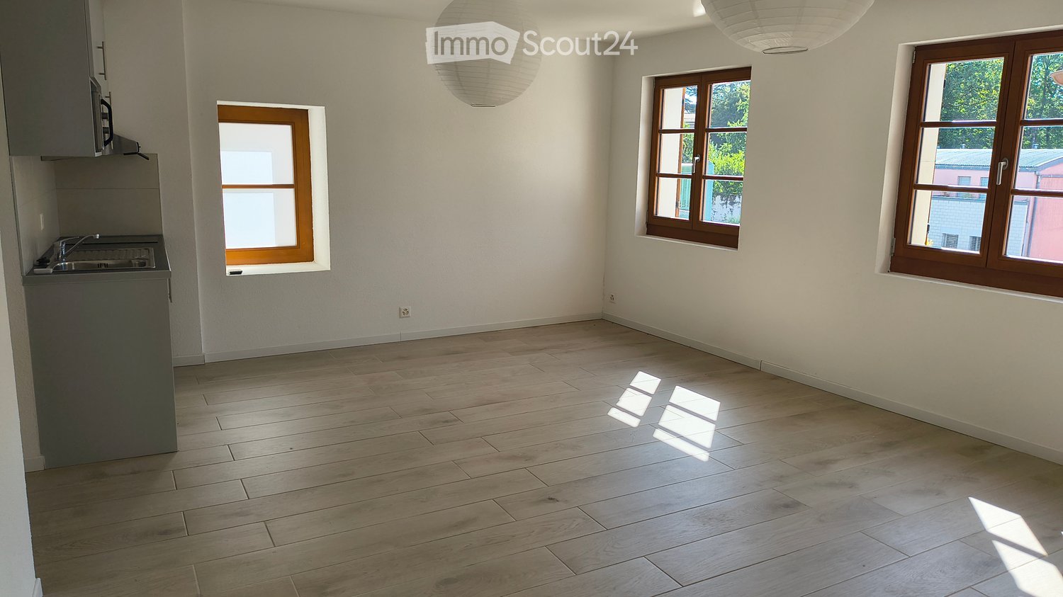 Empty room with light wooden flooring, white walls, and three windows with wooden frames. A small kitchen area is visible on the left side with a sink and countertop.