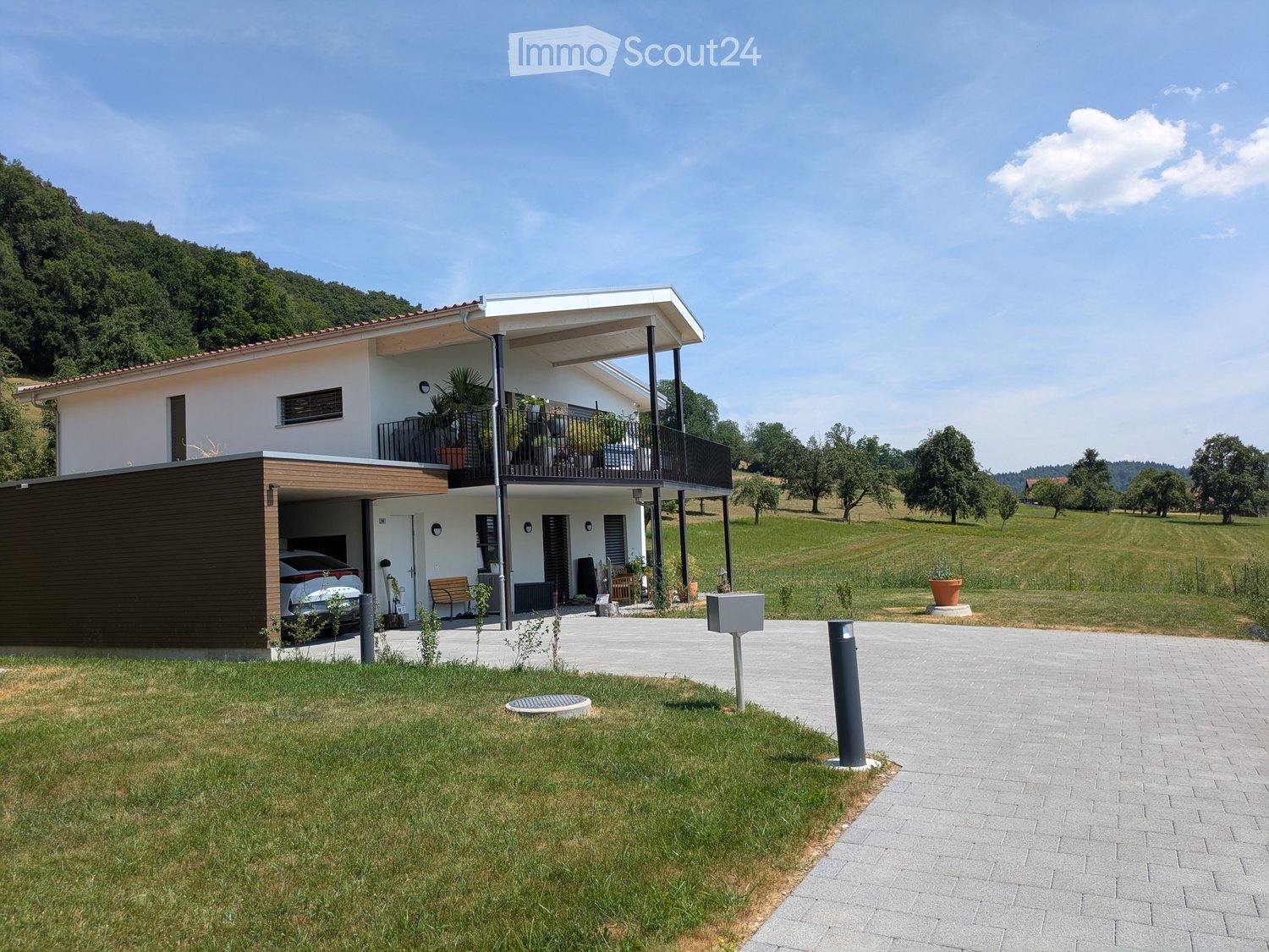 Modern two-story house with white exterior, red-tiled roof, balcony, garage, and surrounding green landscape.