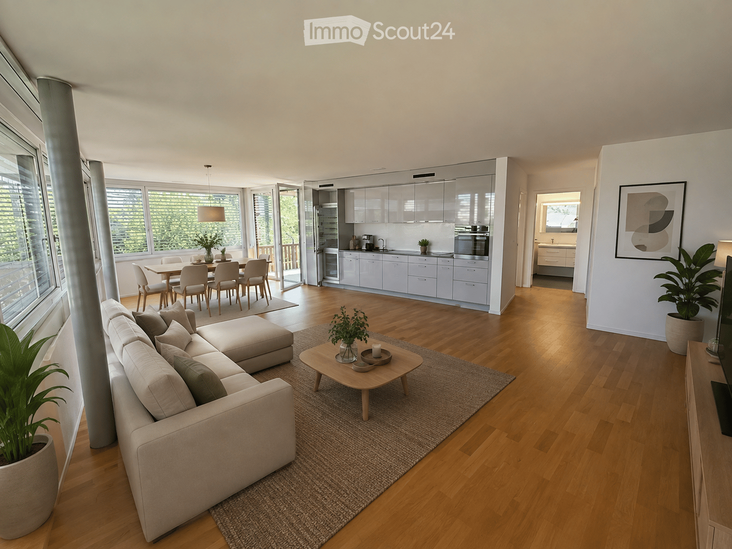 Modern open-plan living and dining area with light wood flooring, large windows, a sectional sofa, coffee table, dining table with chairs, and a sleek kitchen with white cabinets and stainless steel appliances.