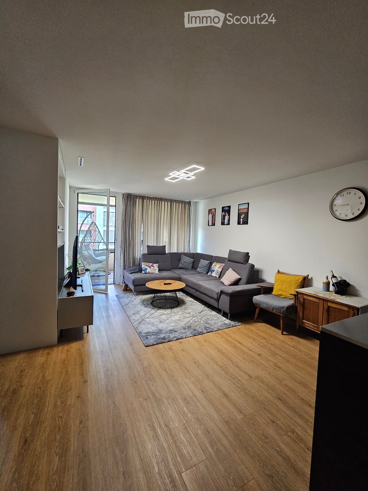 Modern living room with wooden flooring, grey sectional sofa, coffee table, side chair, and a large window with curtains leading to a balcony.
