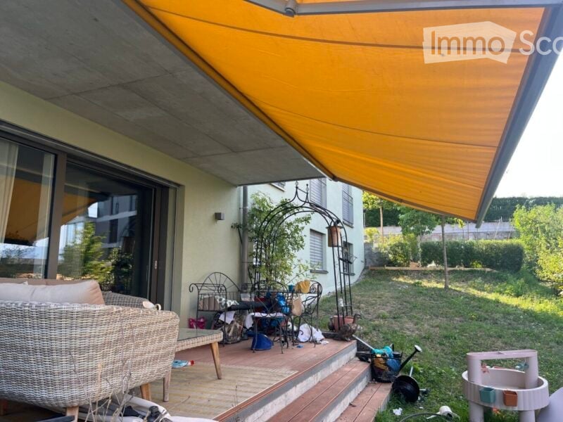 Outdoor patio with a large yellow awning, wicker sofa, metal chairs, and a small table. The area is partially covered and has a view of a grassy yard with some plants and a small decorative arch.