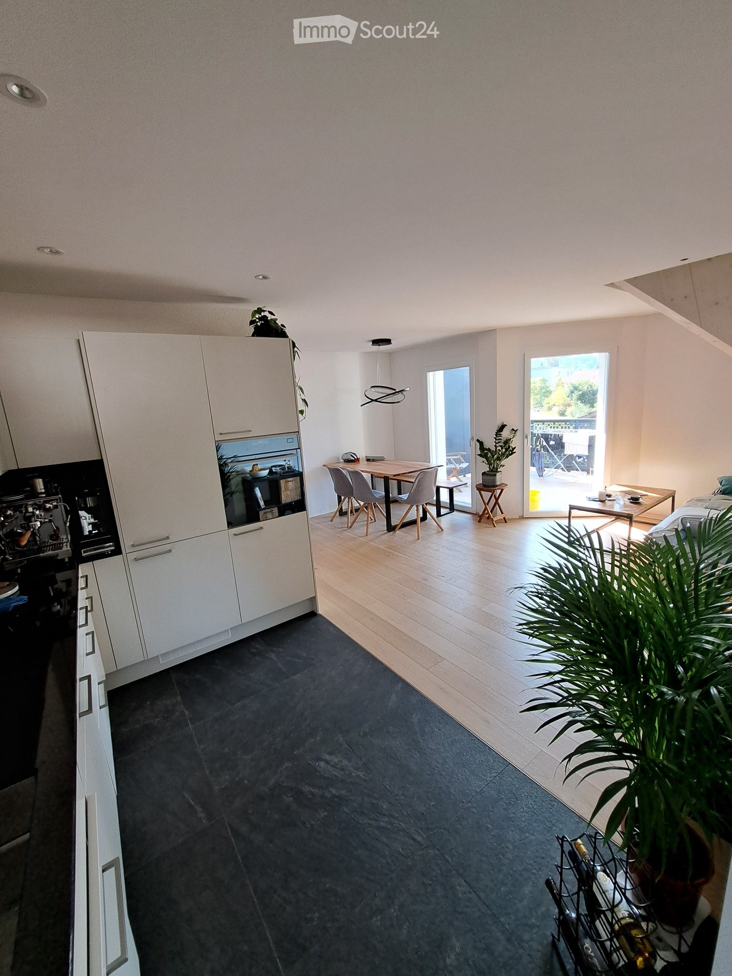 Open-plan living and dining area with modern white kitchen cabinets, a coffee maker, oven, dining table with chairs, a small stool, a sofa, potted plants, and a wine rack.