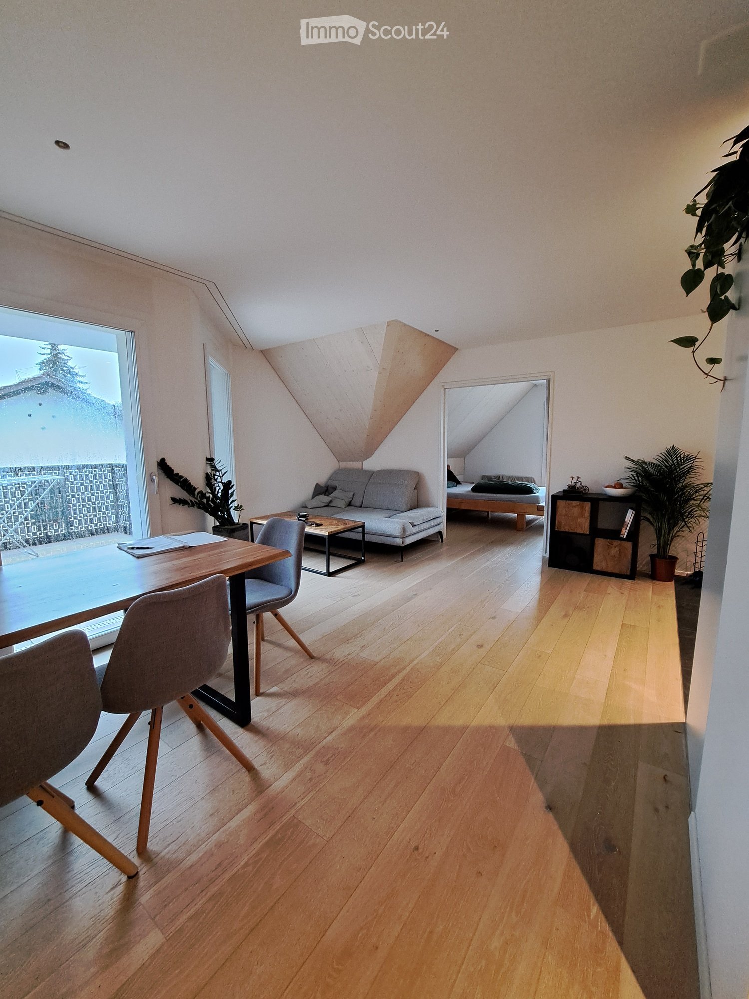 Bright, modern living and dining area with wooden flooring, large windows, a sofa, coffee table, dining table with chairs, and a small bedroom area visible through an open doorway.