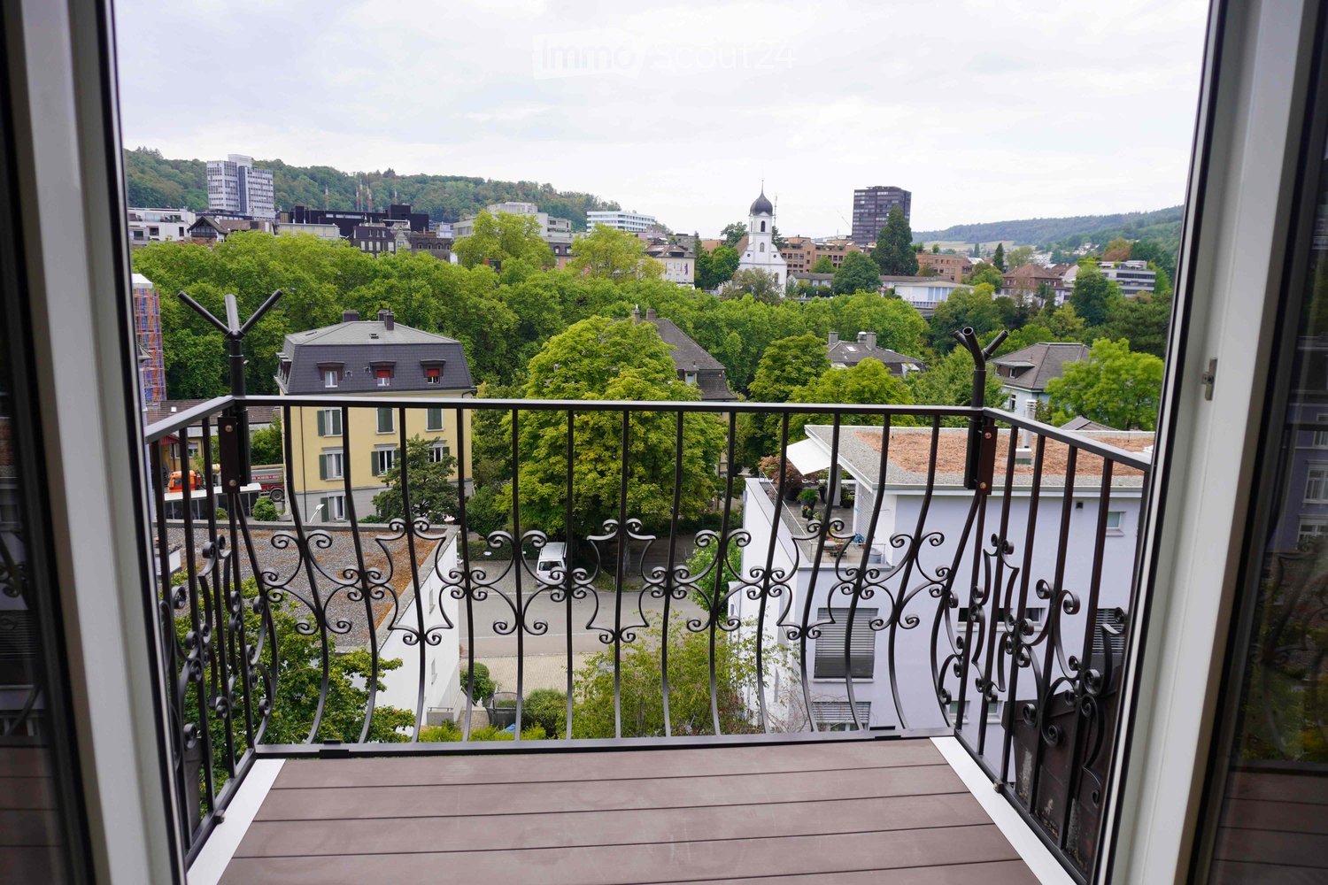 Balcony with wooden decking and black wrought-iron railing, overlooking a cityscape with trees, buildings, and a church spire.