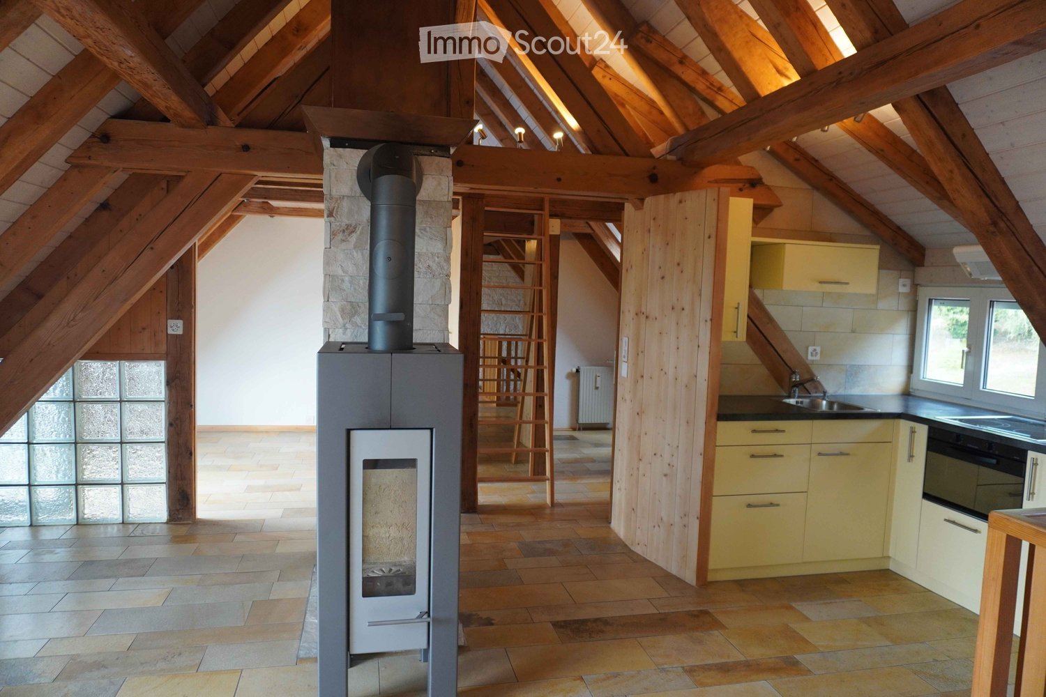 Loft-style room with exposed wooden beams, a modern fireplace, a small kitchen area with cabinets and sink, and a visible staircase leading to another level.