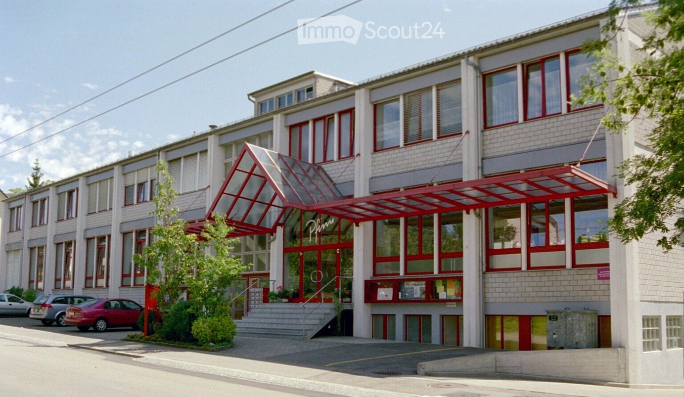 Modern multi-story building with red and white facade, large windows, and a covered entrance with a glass canopy. Several cars are parked in front.