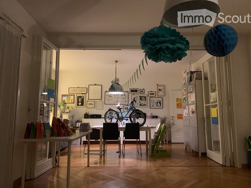 A modern, open-plan living and dining area with wooden flooring, white walls, and large windows. The space features a dining table with chairs, a bicycle, a small cabinet, and several picture frames on the wall. Colorful balloons are hanging from the ceil