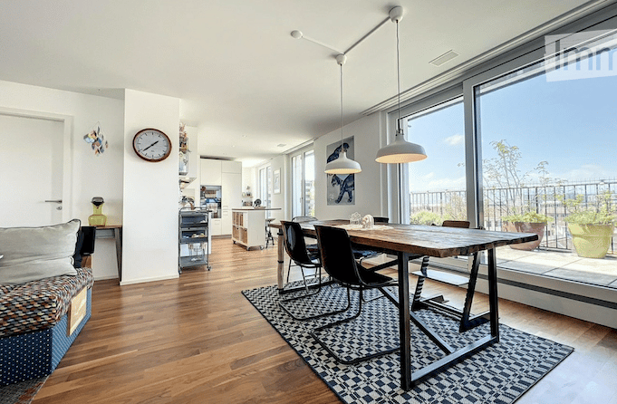 Open-plan living and dining area with hardwood floors, large windows, and a balcony view. Features a sofa, coffee table, dining table with chairs, and a kitchen island.