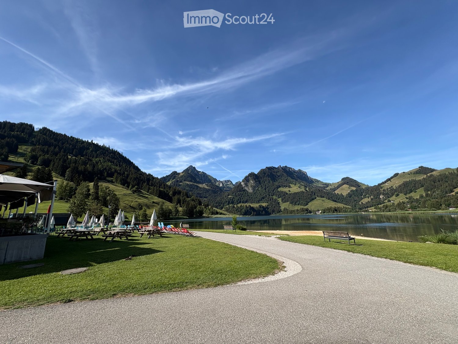 Lakeside park with picnic tables, benches, umbrellas, and lounge chairs, surrounded by green hills and mountains.