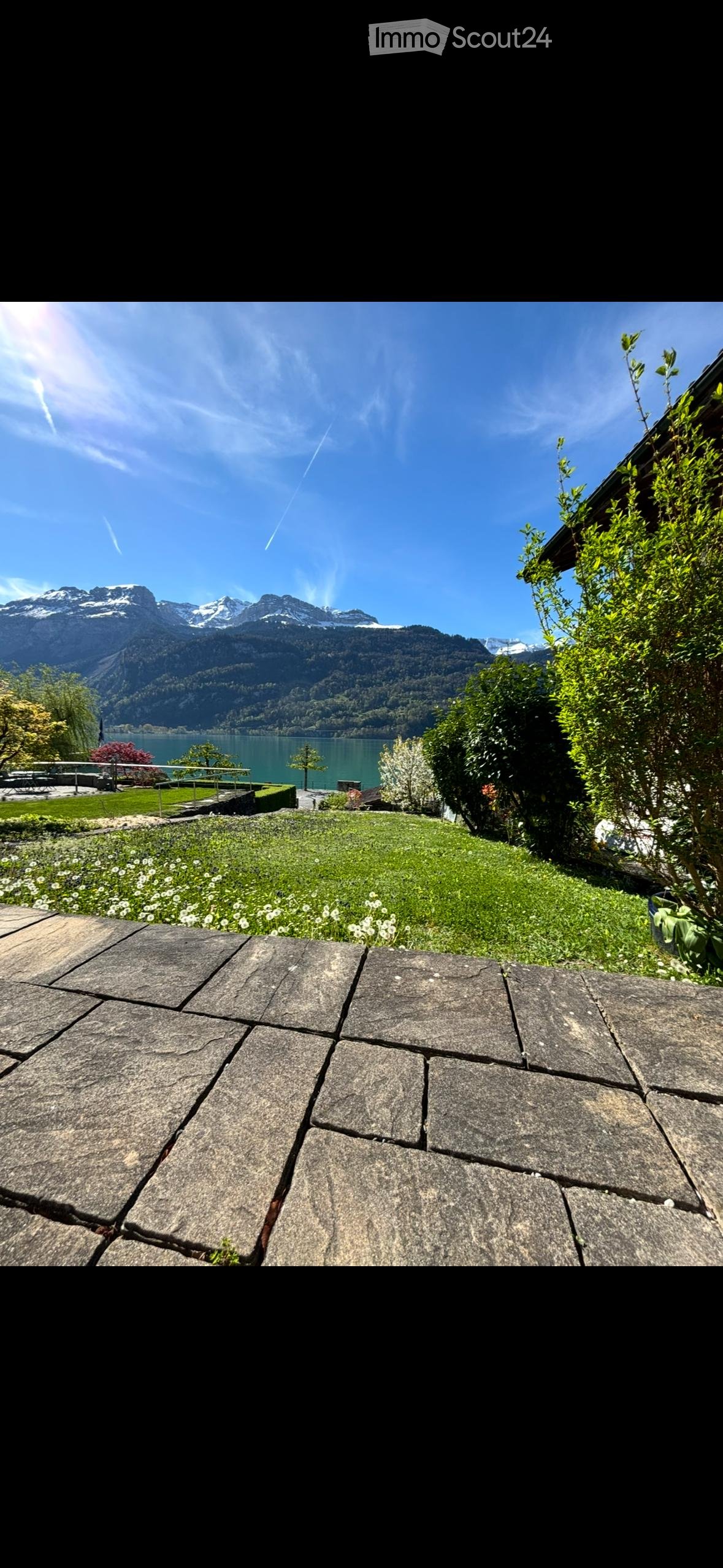 A well-maintained garden with a stone patio, lush greenery, and a scenic mountain and lake view in the background.