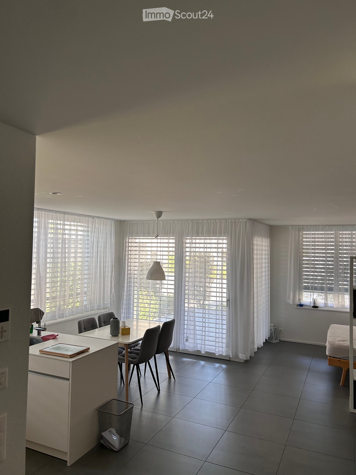 Bright, open room with large windows covered by white curtains, a dining table with chairs, a kitchen island, and a lamp hanging from the ceiling.